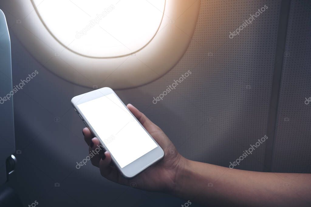 Mockup image of woman's hand holding a white smart phone with blank desktop screen next to an airplane 