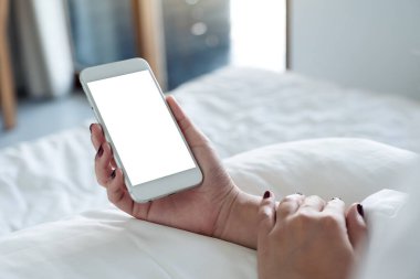 Beyaz yatakta oturan, boş masaüstü ekranlı, beyaz cep telefonlu bir kadının el resmi.