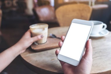 Boş masaüstü ekranlı beyaz cep telefonu ve kafedeki ahşap masada bir bardak kahve tutan ellerin görüntüsü.