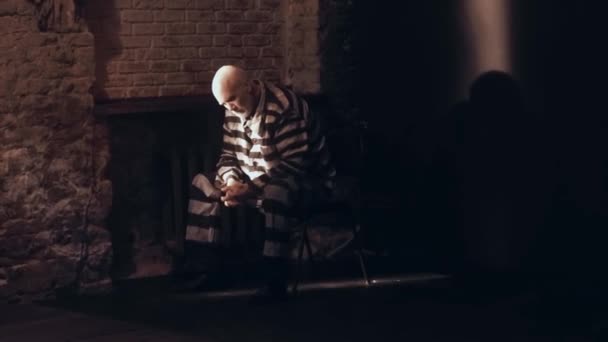 Un anciano se sienta en una sombría celda de la prisión, está vestido con un uniforme de prisión, un hombre de pelo gris está triste, piensa en la vida y planea salir de la cárcel. — Vídeos de Stock