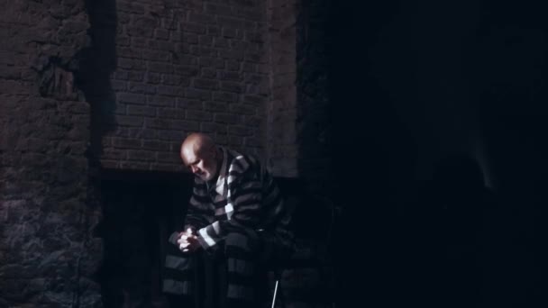 Пожилой человек сидит в мрачной тюремной камере, одет в тюремную форму, седой мужчина грустит, думает о жизни и планирует выйти из тюрьмы — стоковое видео