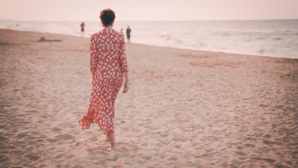 Vista en cámara lenta de una hermosa mujer en vestido rojo caminando por la playa al atardecer — Vídeo de stock