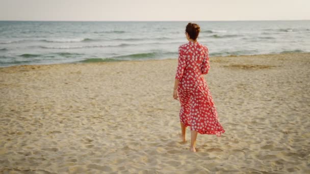 Vista en cámara lenta de una hermosa mujer vestida de rojo caminando por la playa al atardecer — Vídeos de Stock