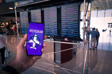 Arka plan olarak havaalanı tarifeleri ile ekranda Flyskam mesajı ile bir akıllı telefon tutma El.