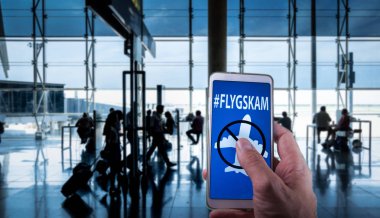 Arka planda havaalanı yolcuları ile ekranda Flyskam mesajı ile bir akıllı telefon tutma El.