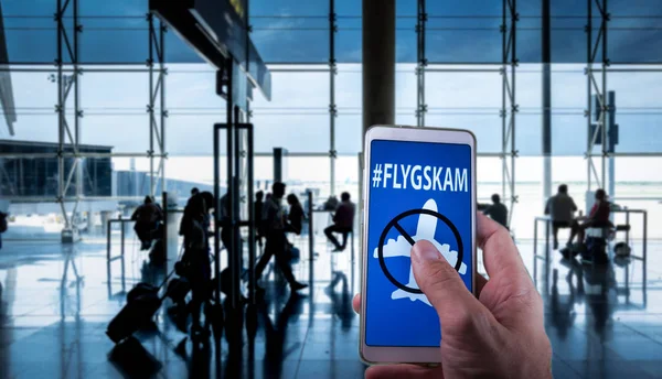 Mano sosteniendo un teléfono inteligente con mensaje Flyskam en la pantalla con los pasajeros del aeropuerto en el fondo . —  Fotos de Stock