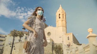 Covid-19 sırasında cerrahi maskeli güzel kıvırcık esmer turist fırında Sitges köyü ile birlikte.