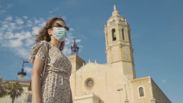 Belle touriste brune bouclée avec un masque chirurgical pendant Covid-19 avec le village méditerranéen de Sitges sur le terrain de boulangerie — Video