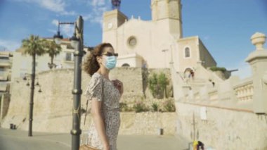 Covid-19 sırasında cerrahi maskeli güzel kıvırcık esmer turist fırında Sitges köyü ile birlikte.