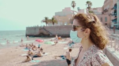 Covid-19 sırasında cerrahi maskeli güzel kıvırcık esmer turist fırında Sitges köyü ile birlikte.