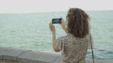 Akıllı telefon kullanan ve Sitges, Barcelona 'daki güzel Akdeniz' in fotoğrafını çeken bir turist kadın..