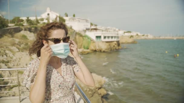 Gyönyörű göndör barna turista felvesz egy sebészeti maszk alatt Covid-19 a mediterrán falu Sitges a pékföldön — Stock videók