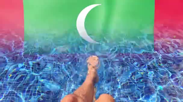 Maldives waving flag and Mans legs resting by the swimming pool edge. — Stock Video