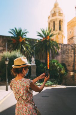 Cerrahi maskeli güzel kıvırcık esmer turist İspanya 'nın Akdeniz köyü Sitges' de selfie çekiyor..