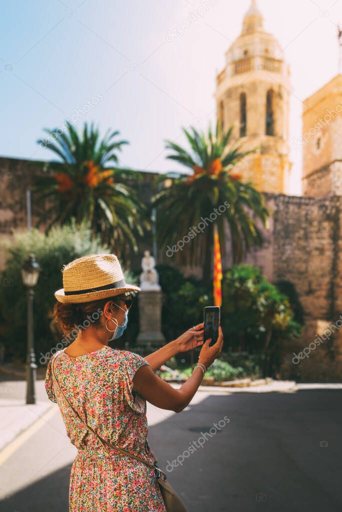 Beautiful curly brunette tourist with a surgical mask taking a selfie in the Mediterranean village of Sitges, spain.