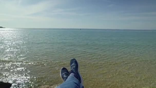 Perspectiva en primera persona disparada desde un excursionista sentado en el borde de un acantilado sobre el mar azul — Vídeo de stock