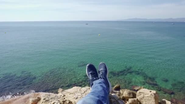 Perspectiva en primera persona disparada desde un excursionista sentado en el borde de un acantilado sobre el mar azul — Vídeo de stock