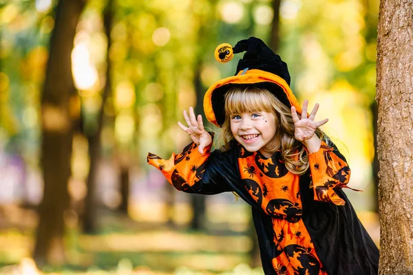 Концепция Хэллоуина Милая маленькая ведьма прячется за деревом в лесу, парк. Красивая девушка в костюме ведьмы с украшением на Хэллоуин . — стоковое фото