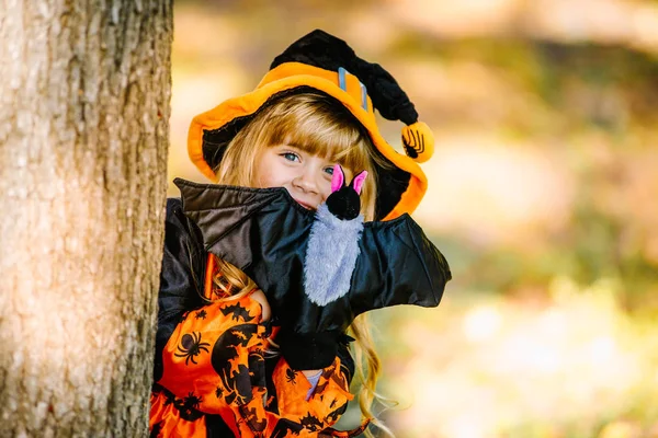 Concept Halloween Mignonne petite sorcière cachée derrière un arbre dans une forêt, parc. Belle fille en costume de sorcière avec décoration d'Halloween . — Photo