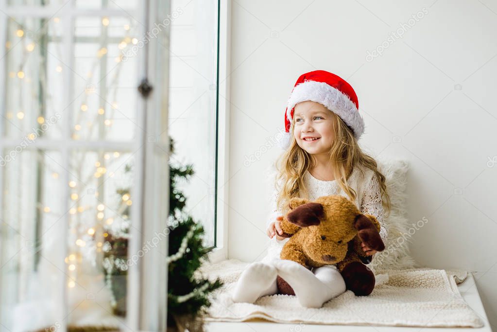 Little girl in the hat of Santa Claus with a plush deer sitting on the window. A child looks out the window and is waiting for Christmas, Santa Claus