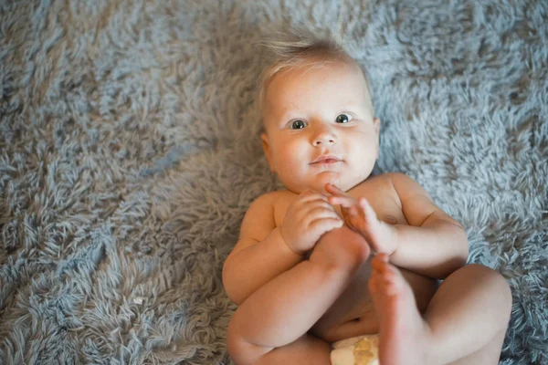 Vicces kis baba visel egy pelenka, játszik egy szürke bolyhos takaró az ágyon. Baba fürdés vagy zuhanyozás után. Pelenka módosítása és bőrápolás. Aranyos baba játék a lábát — Stock Fotó