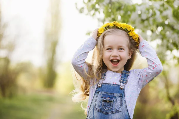 Frühlingssonniges Porträt eines niedlichen 4-jährigen Mädchens, das mit einem Löwenzahnkranz posiert und in die Kamera blickt — Stockfoto