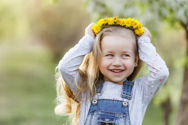 Frühlingssonniges Porträt eines niedlichen 4-jährigen Mädchens, das mit einem Löwenzahnkranz posiert und in die Kamera blickt — Stockfoto