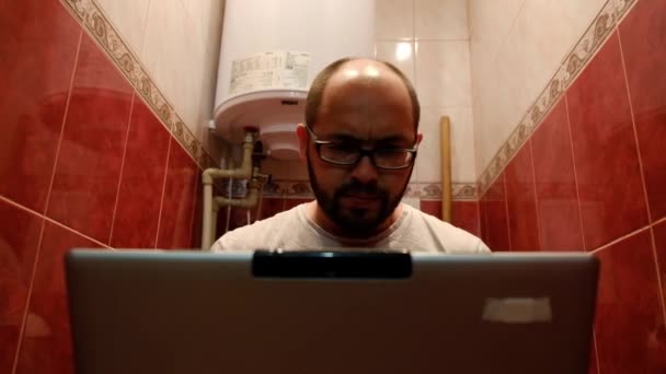 Brodaty człowiek wpisując na laptopie, siedząc na toalecie. — Wideo stockowe