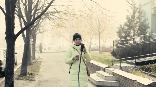 Chica en una chaqueta está caminando por la calle con un vaso de café en sus manos — Vídeos de Stock