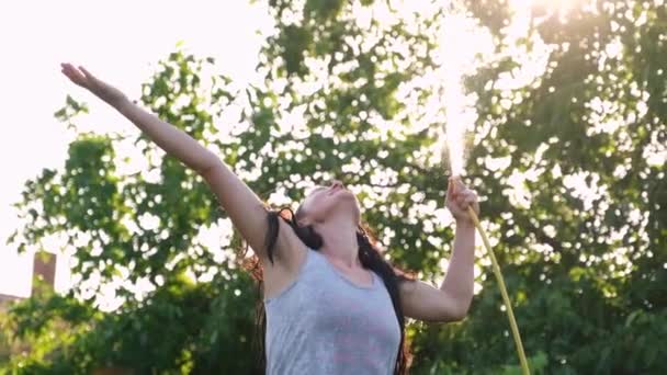 Gelukkig sexy vrouw meisje sproeien met water op een hete zomerdag houden van een slang pijp boven haar hoofd in een straal van warm licht van de zon in de buurt van tuin bomen en planten. Langzame beweging — Stockvideo