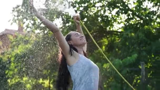 Ευτυχισμένη σέξι γυναίκα που ψεκάζει με νερό μια ζεστή καλοκαιρινή μέρα κρατώντας ένα σωλήνα σωλήνα πάνω από το κεφάλι της σε μια ακτίνα θερμού φωτός από τον ήλιο κοντά σε δέντρα κήπου και φυτά. Αργή κίνηση — Αρχείο Βίντεο
