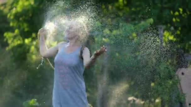 Gelukkig sexy vrouw meisje sproeien met water op een hete zomerdag houden van een slang pijp boven haar hoofd in een straal van warm licht van de zon in de buurt van tuin bomen en planten. Langzame beweging — Stockvideo