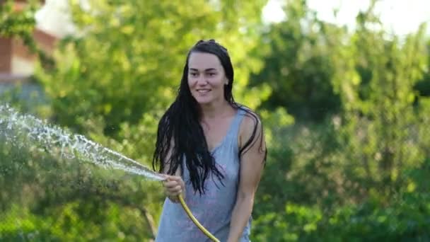 Felice donna sexy ragazza spruzzando con acqua in una calda giornata estiva tenendo un tubo flessibile sopra la testa in un fascio di luce calda dal sole vicino alberi da giardino e piante. Rallentatore — Video Stock