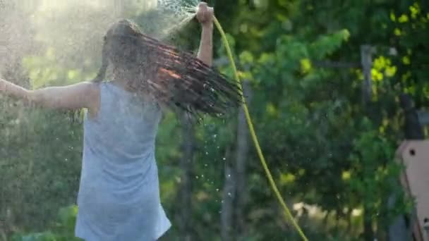 Feliz chica sexy rociando con agua en un día caluroso de verano sosteniendo una tubería de manguera por encima de su cabeza en un haz de luz cálida del sol cerca de árboles y plantas de jardín. Movimiento lento — Vídeos de Stock