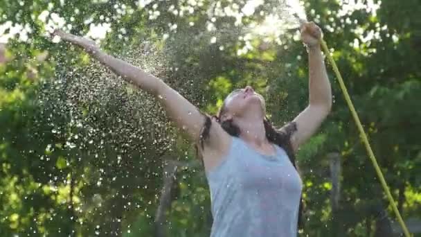 Щаслива сексуальна дівчина розбризкує водою в спекотний літній день, тримаючи шлангову трубу над головою в пучку теплого світла від сонця біля садових дерев і рослин. Повільний рух — стокове відео