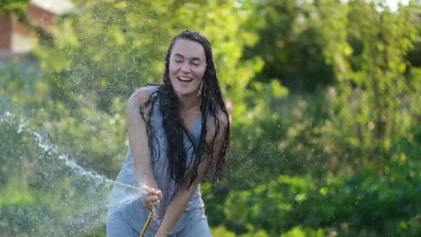 Boldog szexi nő lány permetezés vízzel egy forró nyári napon tartja a tömlő cső a feje felett egy sugár meleg fény a nap közelében kerti fák és növények. Lassú mozgás. — Stock videók