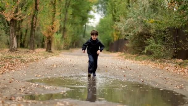 Cámara lenta: un niño activo en botas de goma corre alegremente en un charco en otoño después de la lluvia — Vídeos de Stock