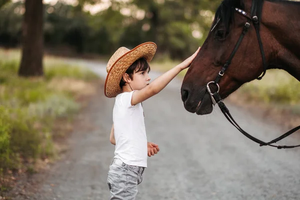 Mignon petit garçon dans un chapeau de cow-boy caressant un beau cheval à l'extérieur — Photo