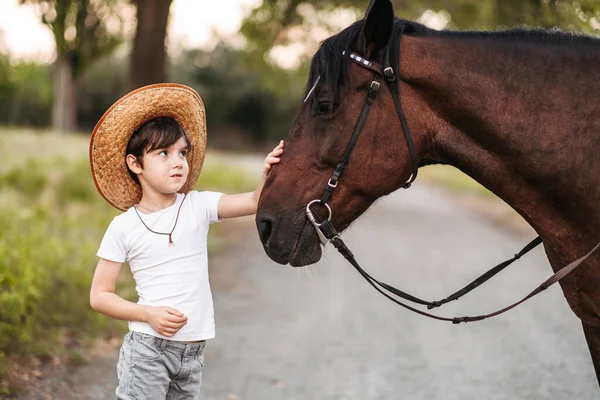 Lindo niño en un sombrero de vaquero acariciando un hermoso caballo al aire libre —  Fotos de Stock