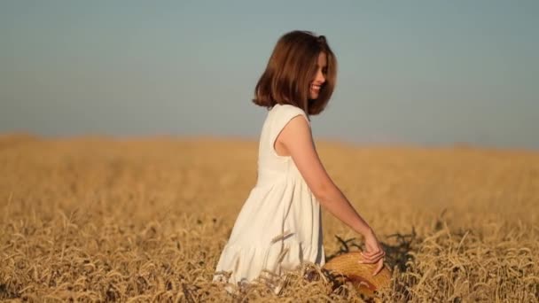 Una joven camina felizmente en cámara lenta a lo largo de un campo amarillo, tocando las espigas de trigo con las manos, levantando su sombrero. Hermosa mujer despreocupada disfrutando de la naturaleza. — Vídeos de Stock