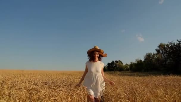 Una joven camina alegremente en cámara lenta a través de un campo amarillo, tocando las orejas con las manos, levantando su sombrero. Hermosa mujer despreocupada disfrutando de la naturaleza. — Vídeos de Stock