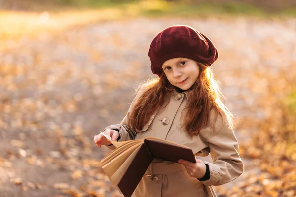 Linda pelirroja niña leyendo un libro en el parque en otoño. Concepto de aprendizaje. Estilo retro. Moda de niños. — Foto de Stock