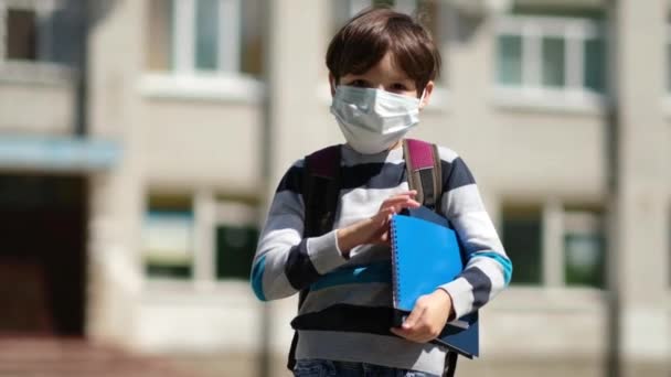 COVID-19 coronavirus πανδημία καραντίνας, πορτρέτο ενός σχολιαρόπαιδου που φοράει προστατευτική μάσκα στην ύπαιθρο. Παιδί αγοράκι με σακίδιο και σημειωματάρια πηγαίνει σχολείο. Εκπαίδευση. Έννοια υγείας. — Αρχείο Βίντεο