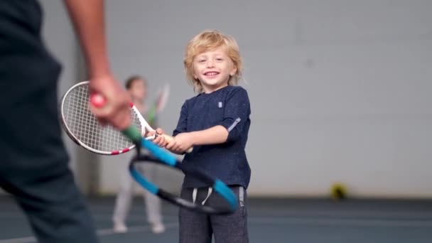 A man coach teaches a little boy to play on an indoor court. A professional tennis instructor throws the ball to hit the racket to the kid, how to return the ball with the racket — Stock Video