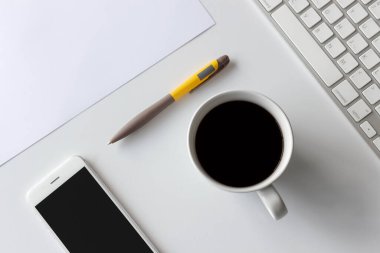 Üst kat ofis masası. Kahve fincanı, akıllı telefon, kalem, klavye ve beyaz arka plandaki diğer ofis ekipmanları.