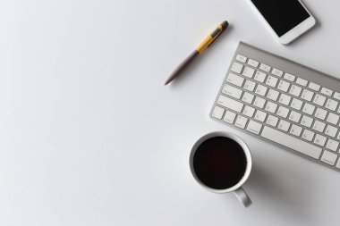 Üst kat ofis masası. Kahve fincanı, akıllı telefon, kalem, klavye ve beyaz arka plandaki diğer ofis ekipmanları.