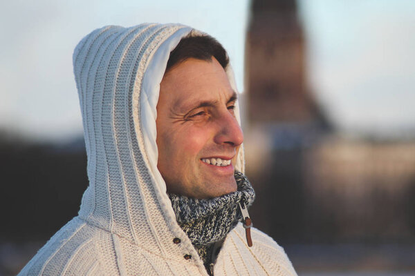 Крупный план портрета мужчины средних лет в белой вязальной куртке с капюшоном в холодную и ветреную погоду, смотрящего вдаль. Зимнее время в Риге, Латвия
