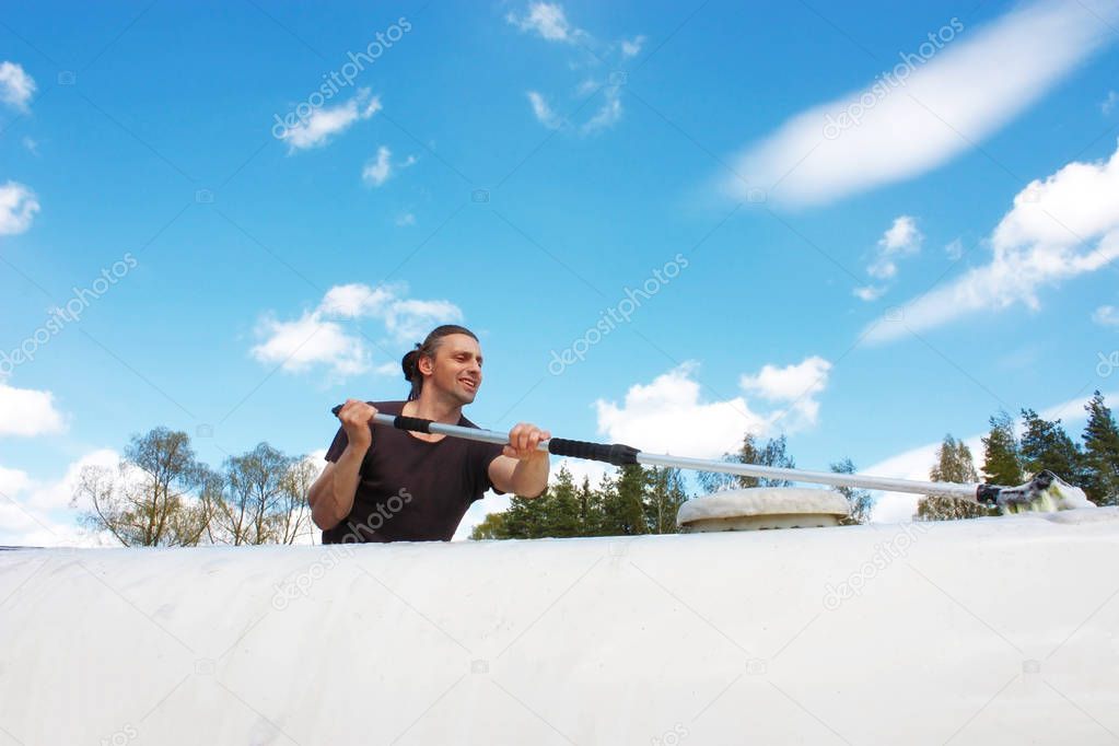 Middle aged man with brown long hairs washing a white caravan. Isolated on blue cloudy sky. Travel concept.