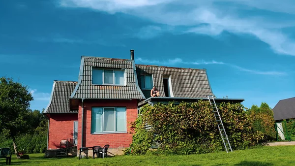 Roof installation by man on the roof of the house. Isolated on the blue cloudy sky. Home and garden repair work in summer time. View from afar.