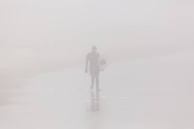 Deal, New Jersey - 28 Nisan 2018: Bir sörfçü çok sisli bir bahar sabahı plaj boyunca yürüyor
